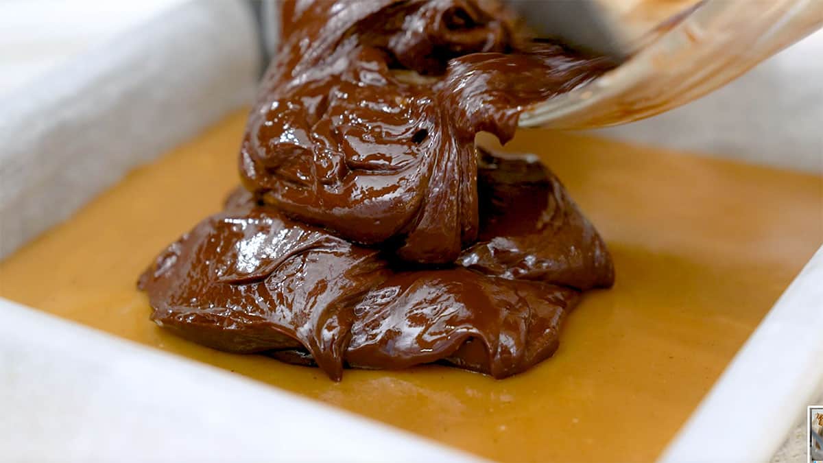 chocolate ganache being spooned on top of a layer of caramel in a baking pan